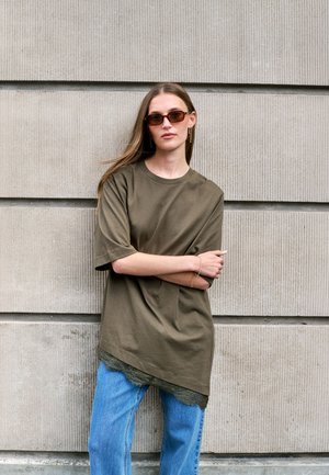 Femme aux cheveux longs portant des lunettes de soleil marron, une chemise oversize vert olive avec un ourlet en dentelle et un jean bleu, debout contre un mur en béton texturé.