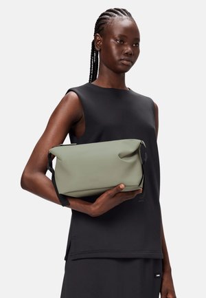 Borsa impermeabile verde con texture liscia, design a rotolo in alto e tracolla nera regolabile, tenuta contro una maglia nera senza maniche.