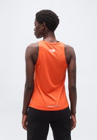 Άτομο με κοντά μαλλιά φορώντας πορτοκαλί αμάνικη μπλούζα και μαύρο παντελόνι, που στέκεται με την πλάτη στην κάμερα μπροστά σε απλό φόντο.