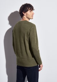 Olivgrön långärmad skjorta i strukturerat tyg, med rundad halsringning och avslappnad passform. Modellen visar bakifrån.