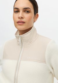 Kurtka polarowa z blokami kolorów w odcieniach beżu i kremu. Wysoki kołnierz, zapięcie na suwak oraz logo na piersi. Miękki, teksturowany materiał.