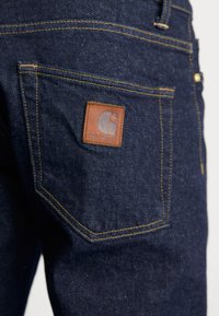 Dark blue denim jeans with a tan leather patch featuring a logo, yellow stitching, and a right back pocket with decorative seam detailing.