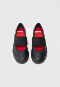 Zapatos de cuero negro con forma redondeada, que cuentan con una amplia banda elástica negra en la parte superior y una plantilla roja con el logo "CAMPER".