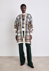 Poncho tricotat cu un model în bej, bleu închis și portocaliu, cu un finisaj texturat; asortat cu pantaloni verzi închiși și pantofi maro.