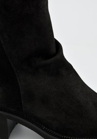 Bottine en suede noir avec une texture lisse, un design légèrement affaissé et un petit talon. Présente des coutures détaillées le long de la semelle.