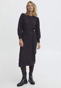 Donkergrijze gebreide jurk met lange, geribbelde mouwen, een tailleband met strik en een rok tot de knie. Draagt een paar zwarte enkellaarzen.