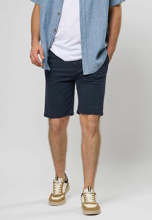 Marineblaue Shorts aus einer Baumwollmischung mit Seitentaschen und geradem Schnitt, kombiniert mit einem hellblauen Hemd mit Knöpfen und beigen Sneakers.