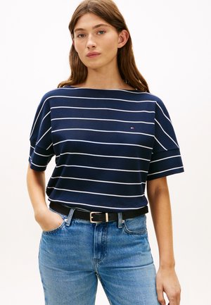 Moteris dėvi tamsiai mėlynį trumpomis rankovėmis marškinėlį su baltais horizontalais dryžiais ir mažą logotipą, įkištą į šviesiai mėlynas džinsines kelnes su juodu diržu.