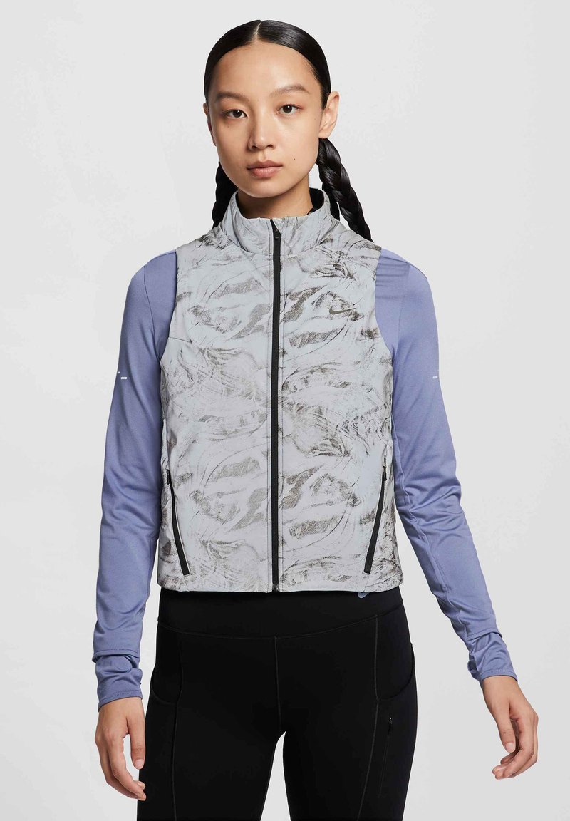 Gilet leggero senza maniche con un motivo astratto grigio, zip nera e tasche laterali, indossato sopra una maglia blu a maniche lunghe.