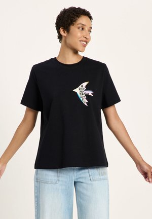 Person trägt ein schwarzes T-Shirt mit einem bunten Vogelgrafik und den Worten "Be bold and proud and wonderful.". Hellblaue Jeans mit Applikationsdetails.