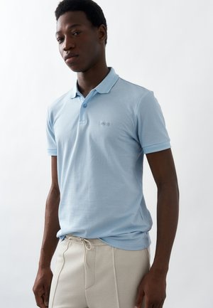 Άνδρας που φοράει ανοιχτό μπλε πόλο με κοντά μανίκια και το λογότυπο "BOSS" και μπεζ παντελόνι με κορδόνι, στέκεται μπροστά σε απλό λευκό φόντο.