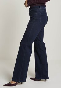 Kvinna som bär mörkblå vida jeans och burgundy spetsiga klackskor, stående med armarna i kors mot en neutral bakgrund.