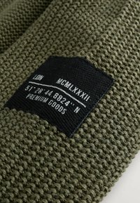 Tissu texturé vert olive avec une étiquette noire comportant des textes blancs : "LON", "MCMLXXIII" et les coordonnées "51°28'44.8824'' N." Produits de luxe.