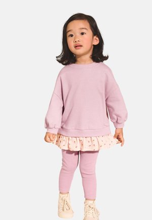Peutermeisje met donker haar tot op de schouders, draag een roze oversized trui, een bloemenrok, roze legging en crèmekleurige hoge sneakers.