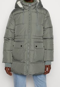 Chaqueta gris acolchada con cierre de cremallera frontal, capucha y cordones. Cuenta con dos bolsillos frontales y puños acanalados. Forrada con tela suave.