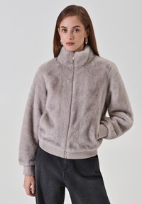 Giacca in pelliccia sintetica di colore grigio chiaro con colletto alto, chiusura con zip e polsini e orlo a costine. Presenta tasche laterali e una texture morbida e pelosa.