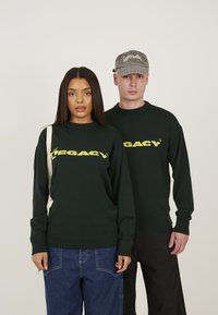 Zwei Personen tragen dunkelgrüne Pullover mit dem Aufdruck "LEGACY" in Gelb. Die Pullover haben gerippte Bündchen und Säume. Eine Person hält eine beigefarbene Tasche.
