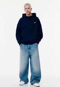 Felpa con cappuccio blu marina e tasca frontale, abbinata a jeans a gamba larga di colore azzurro chiaro. Il modello indossa scarpe da ginnastica bianche, con uno sfondo semplice.