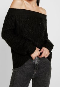 Pull noir en tricot, épaules dénudées, surface texturée, encolure large côtelée et coupe ample. Associé à un jean denim foncé.