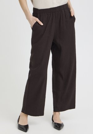 Woman wearing loose black pants with elastic waistband and black pointed flats, hands in pockets, standing against plain background.