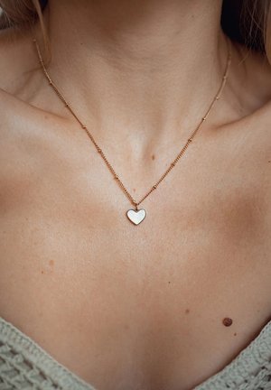 Collana a catena color oro con un pendente a forma di cuore argentato lucido, appoggiato sulla pelle nuda. La vista ravvicinata evidenzia la texture e la forma.