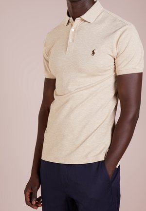 Polo beige en tissu doux, avec col, manches courtes, trois boutons et un logo brodé sur le côté gauche de la poitrine.