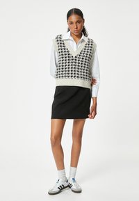 Mustavalkoinen houndstooth-kuvioinen neuleliivi valkoisen kauluspaitansa päällä, yhdistettynä mustaan hameeseen, polven yli ulottuviin valkoisiin sukkiin ja lenkkareihin.