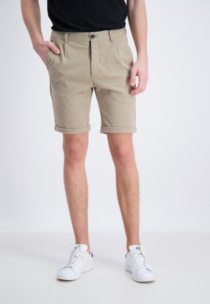 Shorts beiges avec ourlets roulés, fabriqués dans un tissu doux. Présentent une fermeture à bouton à l'avant et deux poches latérales. Associés à des baskets blanches.