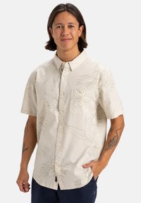 Camicia button-up a maniche corte di un leggero beige con un sottile motivo floreale, tasca sul petto singola e vestibilità rilassata. Realizzata in tessuto morbido.