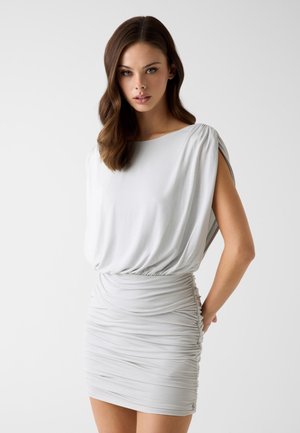 Młoda kobieta o falujących brązowych włosach ubrana w dopasowaną, marszczoną białą sukienkę bez rękawów na jasnym, jednolitym tle.