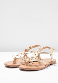 Sandali metallici dorati con design a strisce incrociate, suola piatta e fibbia regolabile alla caviglia. Sottopiede in pelle liscia e fondo beige chiaro.