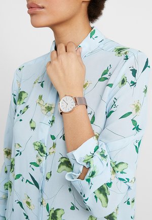 Montre ronde avec un cadran blanc et des accents en or rose, montée sur un bracelet en maille fin. Portée au poignet, associée à une chemise florale bleu clair.