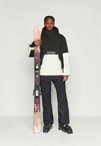 Personne en veste et pantalon de ski noir et blanc tenant une paire de skis rose et noir, chaussée de bottes de ski noires.
