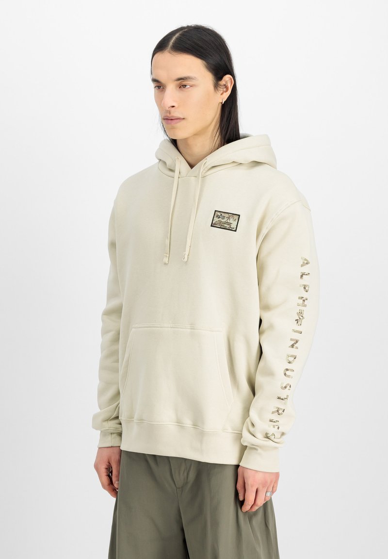 Ljusbeige hoodie med en känguruficka. Har ett märke på bröstet och "ALPHA INDUSTRIES" tryckt längs ärmen i en kontrasterande färg.