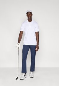 Λευκό πόλο μπλουζάκι με κοντό μανίκι, σκούρο μπλε παντελόνι, λευκό καπέλο γκολφ, γάντι γκολφ και παπούτσια. Στέκεται με ένα ρόπαλο γκολφ και ένα σίδερο γκολφ.