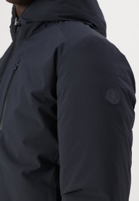 Chaqueta impermeable de color azul marino con capucha, que cuenta con un bolsillo en el pecho con cremallera y un parche circular con logo en el brazo izquierdo. Textura suave.