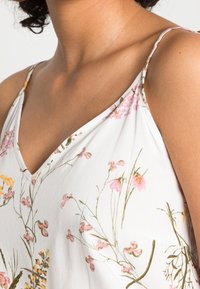 Vit blommig linne med V-ringning, tunna axelband och tryck av gula och rosa blommor med gröna stjälkar. Mjuk tygtextur.