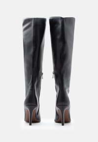 Ζευγάρι μαύρες δερμάτινες μπότες μέχρι το γόνατο με τακούνια στιλέτο, εμφανιζόμενες από πίσω πάνω σε λευκό φόντο.