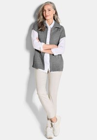 Gilet en tricot gris avec un col sur une chemise blanche à boutons, associé à un pantalon beige clair et des chaussures de sport blanches. Fond neutre.