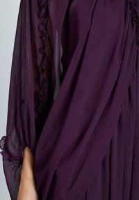 Aksamitny strój w kolorze śliwkowym z lekkim, zwiewnym materiałem, ozdobiony falbankami wzdłuż rękawów i bocznych szwów, o fakturze i lekkości.