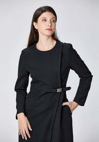 Robe noire ajustée avec manches longues, col rond, poches latérales et une ceinture accentuée à la taille. Fabriquée en tissu lisse et structuré.