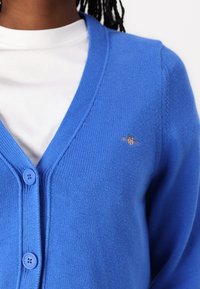 Blue knitted cardigan with a V-neck, two buttons, and a small logo on the chest. Features ribbed detailing along the edges and sleeves.