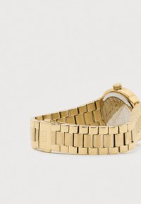 Kuldmetallist link-käekell, millel on kokkuvolditav klamber ja ümmargune korpus, mille tagaküljel on graveeritud brändi üksikasjad.