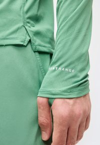 Χέρι που φοράει πράσινο πουκάμισο με μακριά μανίκια και οπή για τον αντίχειρα, με το κείμενο «NIGHT RANGE» στο μανίκι, δίπλα σε ταιριαστό πράσινο παντελόνι.