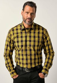 Mann mit Bart, der ein gelb-schwarzes kariertes Hemd mit Knöpfen, einen schwarzen Gürtel, schwarze Hosen und eine Armbanduhr trägt und mit den Händen in den Taschen steht.