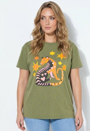 Donna con lunghi capelli biondi che indossa una maglietta verde oliva con una grafica colorata e stilizzata di un gatto e una stella sul davanti.