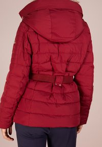 Punainen tikattu takki, jossa on huppu, vyötärövyö ja toppaustekstuuri. Takissa on sileä pinta ja kultainen koriste hihoissa.