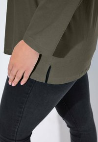 Olive green long-sleeve top with a straight cut, featuring side slits at the hem, paired with black fitted jeans. Soft fabric texture.