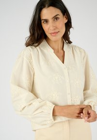 Blusa color crema con ricamo floreale, chiusura frontale con bottoni e maniche a palloncino. Realizzata in tessuto leggero con dettagli testurizzati.