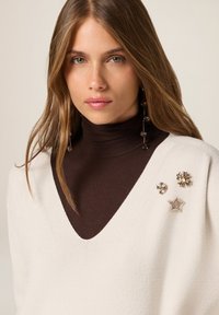 Weißer V-Ausschnitt-Pullover mit strukturiertem Strick, über einem braunen Rollkragenpullover getragen. Verfügt über drei dekorative Broschen: zwei blumenförmige und eine sternförmige.